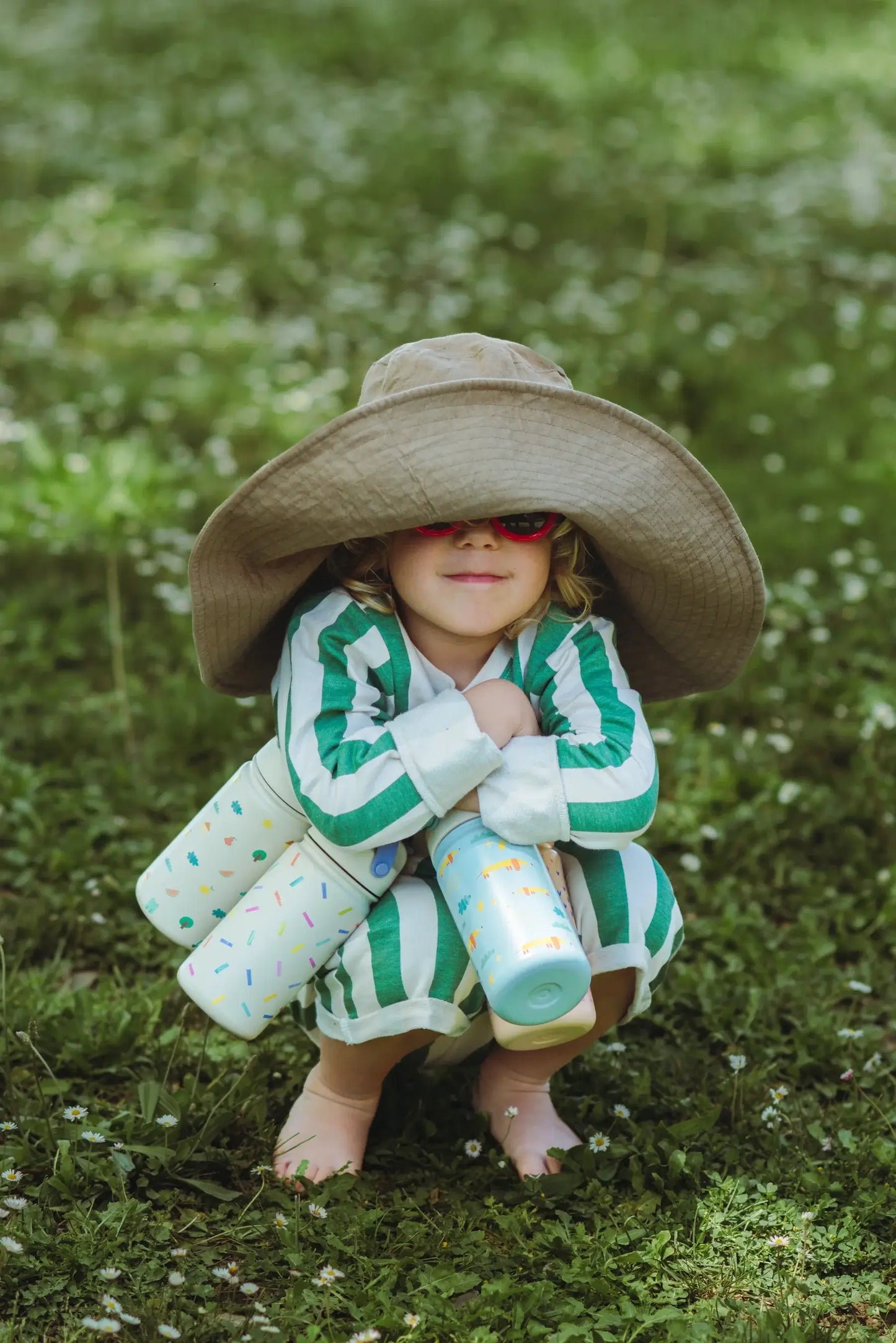Child in a sun hat holding colorful stainless steel drink bottles in a grassy outdoor setting.