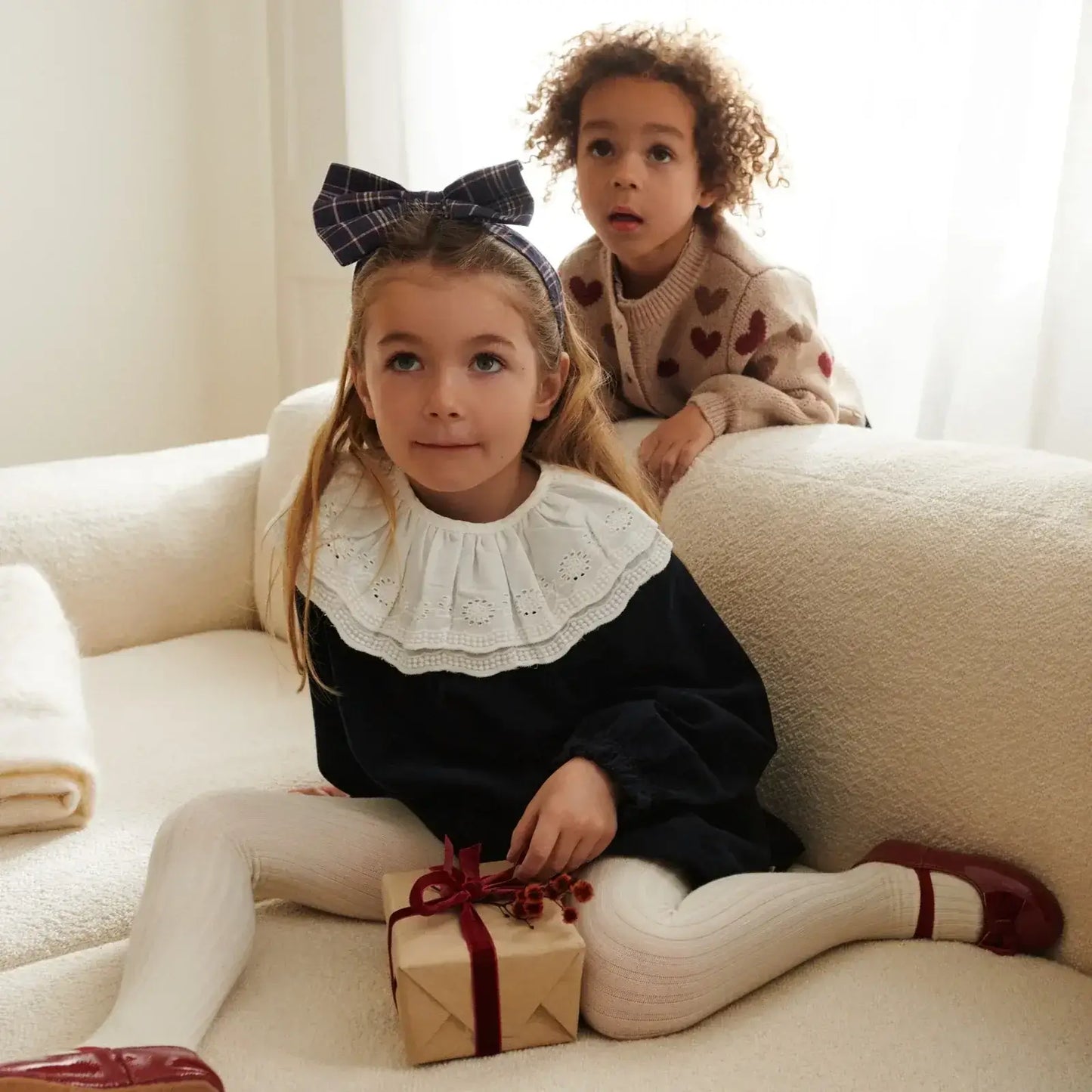 Two children in cozy outfits, one holding a gift, showcasing playful fashion and warmth.
