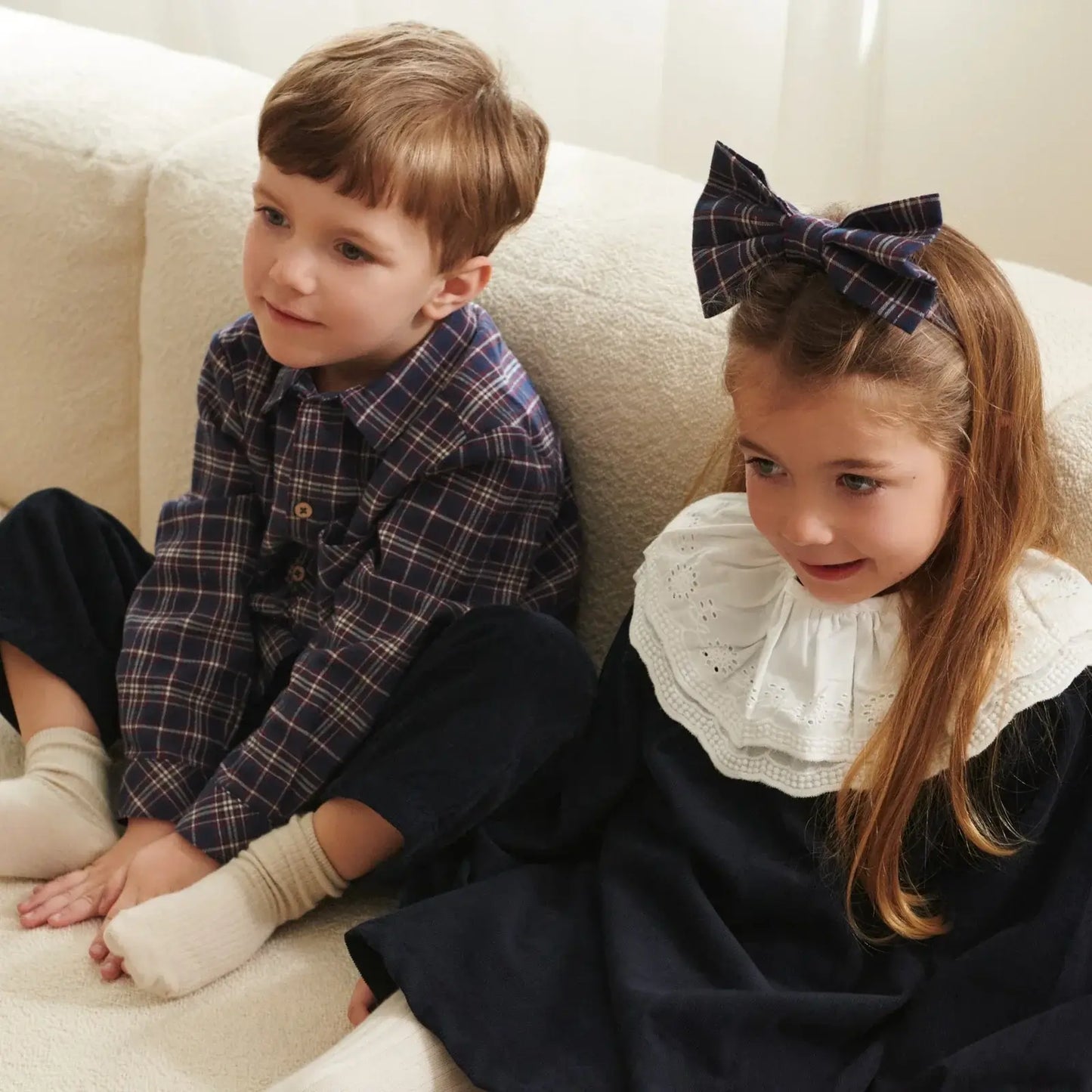 Two children sitting on a sofa, one in a plaid shirt and the other in a navy dress with a white collar and hair bow.