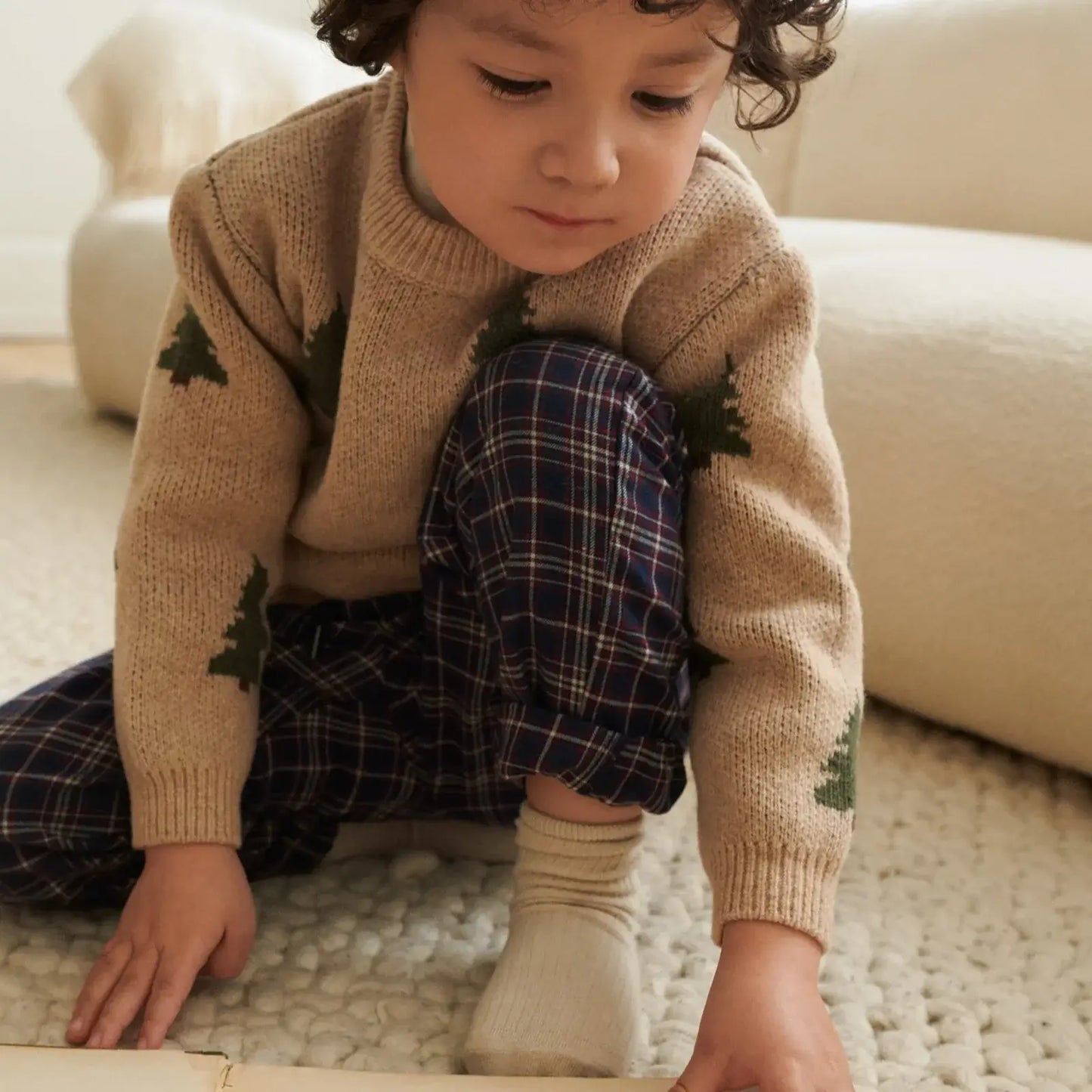 Kind in een zachte, gebreide trui met bomen en pyjamabroek, spelend op een cozy vloer.