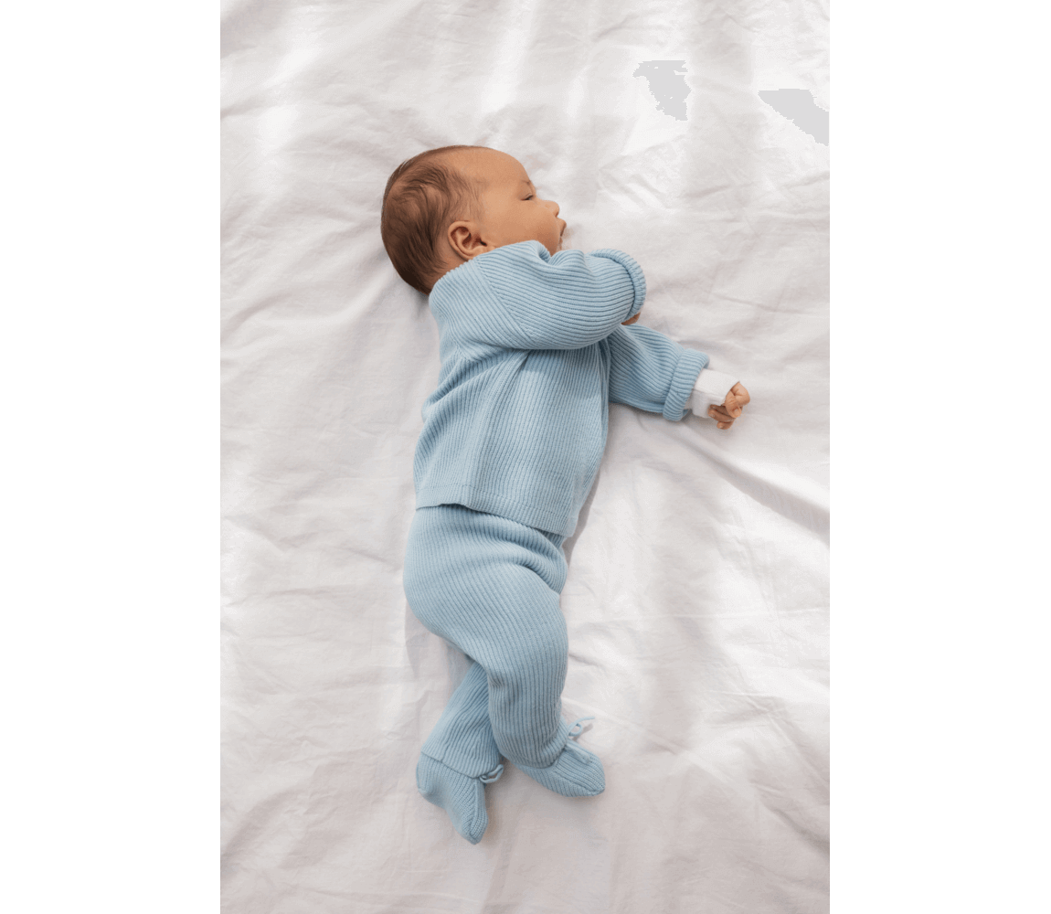 Newborn baby in light blue knitted outfit, featuring elastic waist, on a soft white bedding surface. 100% organic cotton.