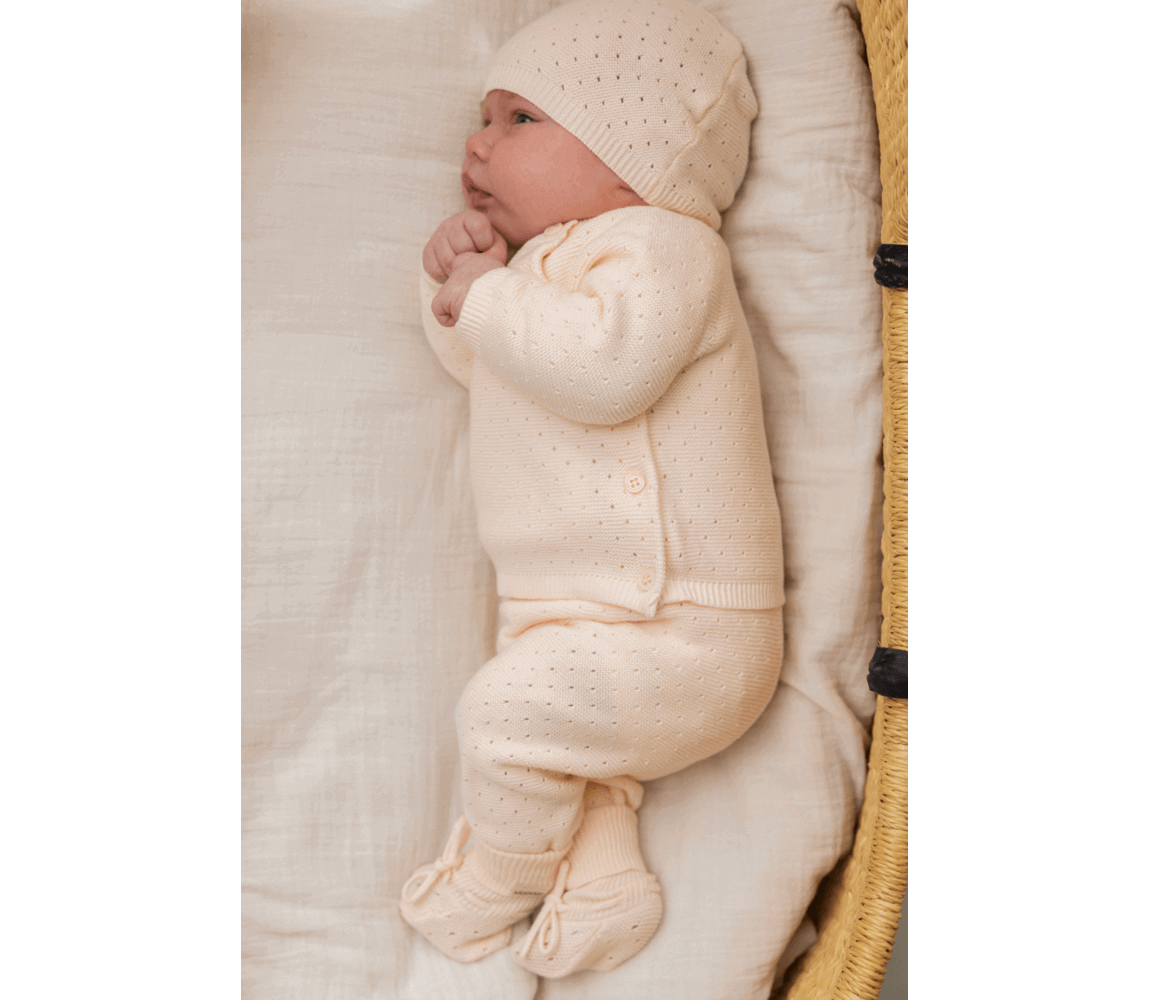 Newborn in soft beige knitted outfit with hat, lying in a basket on a white blanket.