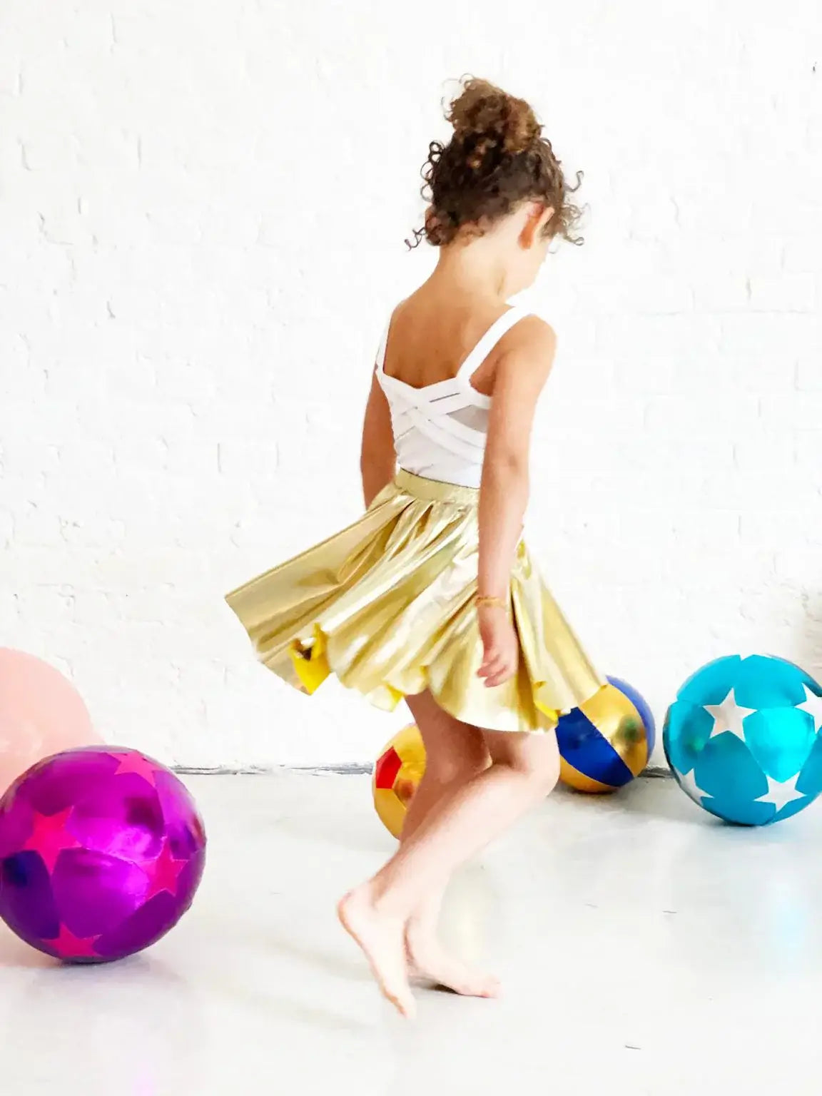 Child in a shiny gold skirt twirls playfully near colorful balls, showcasing fun and creativity in playtime.