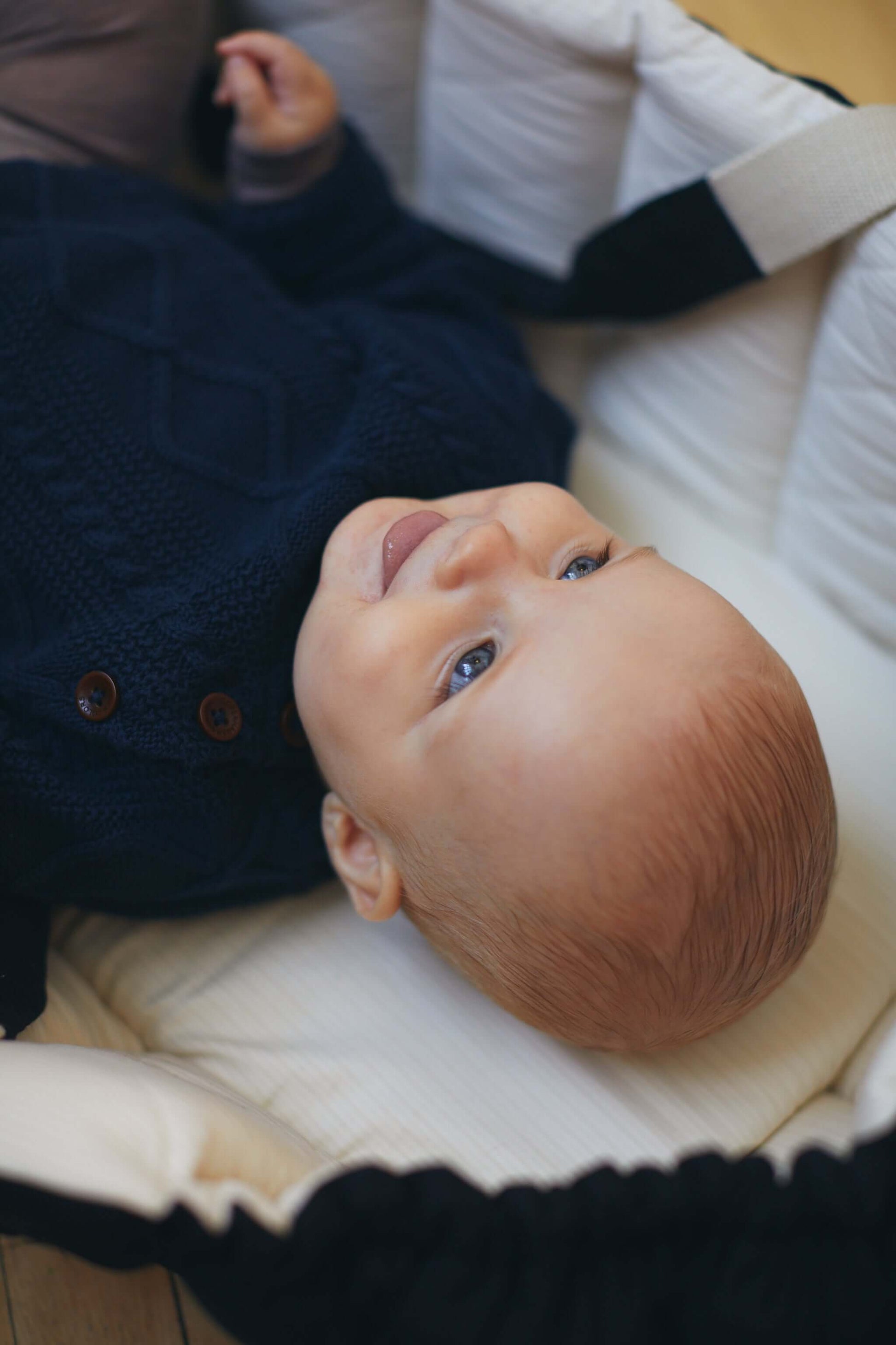 Baby in a navy blue knitted cable sweater lying in a cozy bassinet, looking up with a smile.