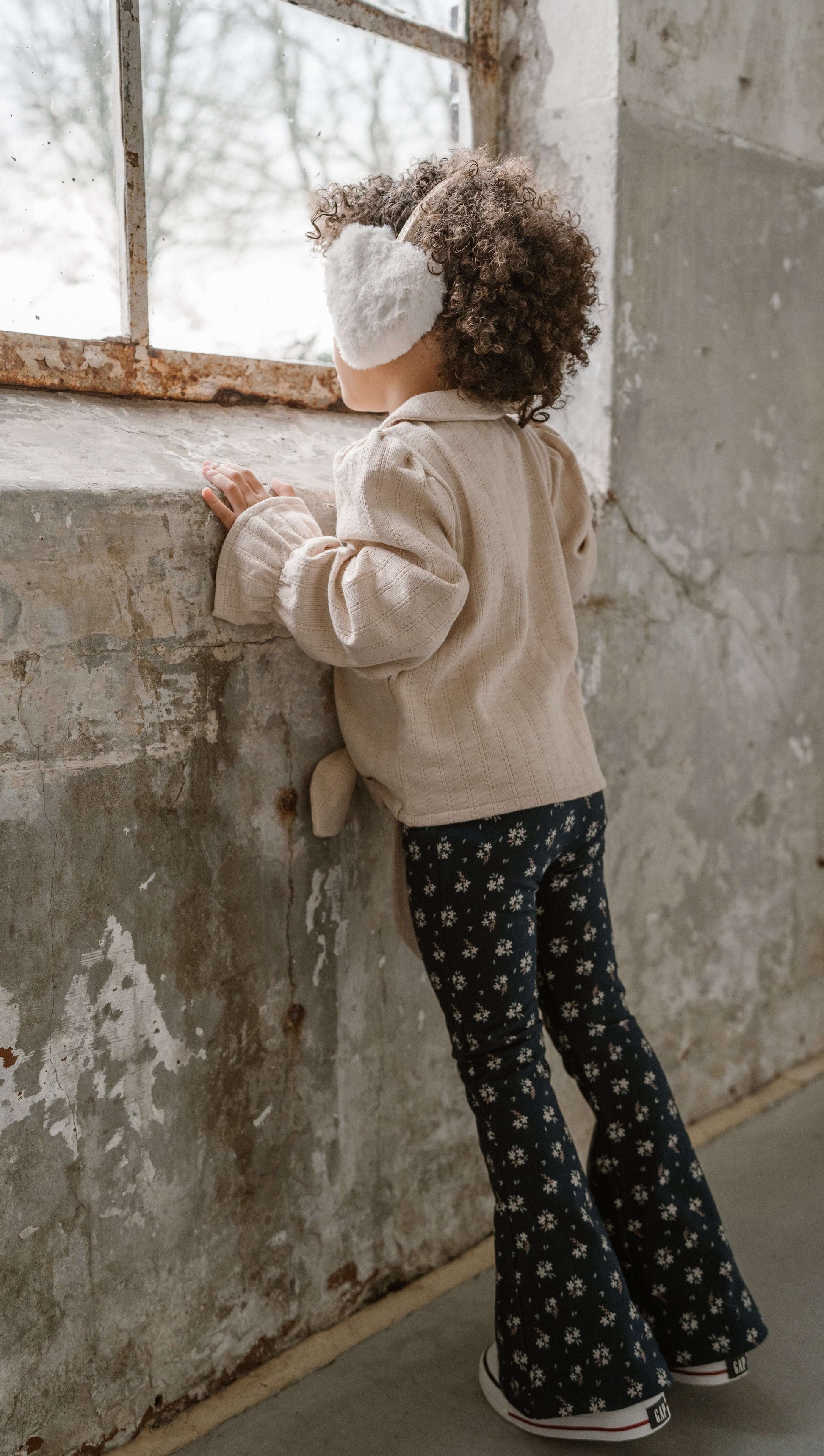 Child with curly hair gazing out a window, wearing a cozy sweater and navy floral flared pants.