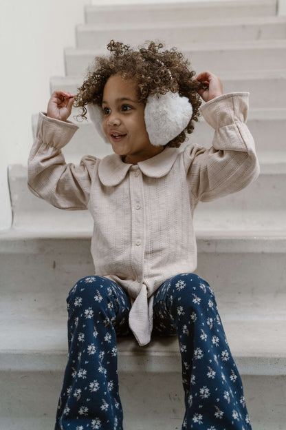 Happy child wearing cozy earmuffs and a button-up shirt, sitting on stairs in stylish dark blue floral pants.