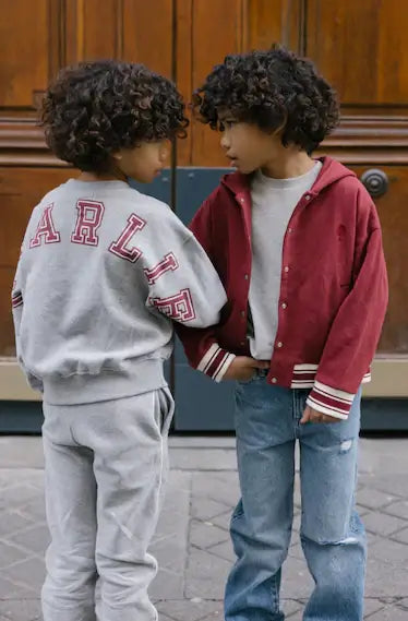 Two children with curly hair in stylish grey and red outfits, showcasing the trendy Charlie Petite Jones sweater and jacket.